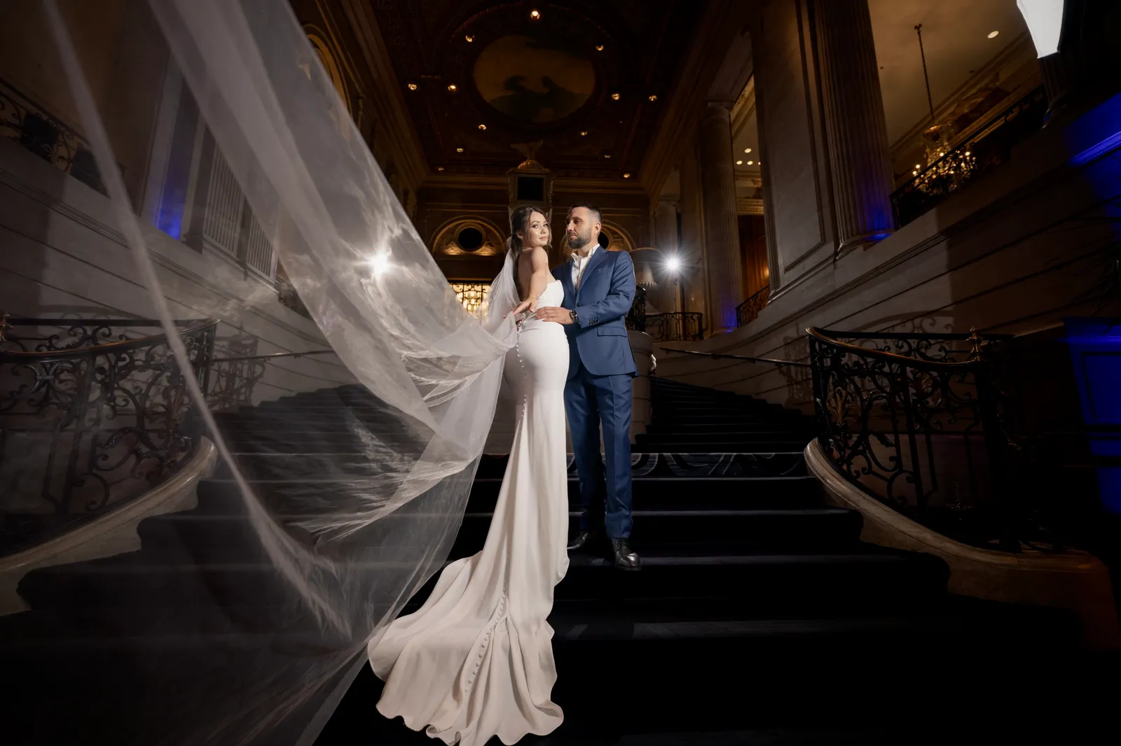 Bride and groom on a grand staircase, her cathedral veil flowing